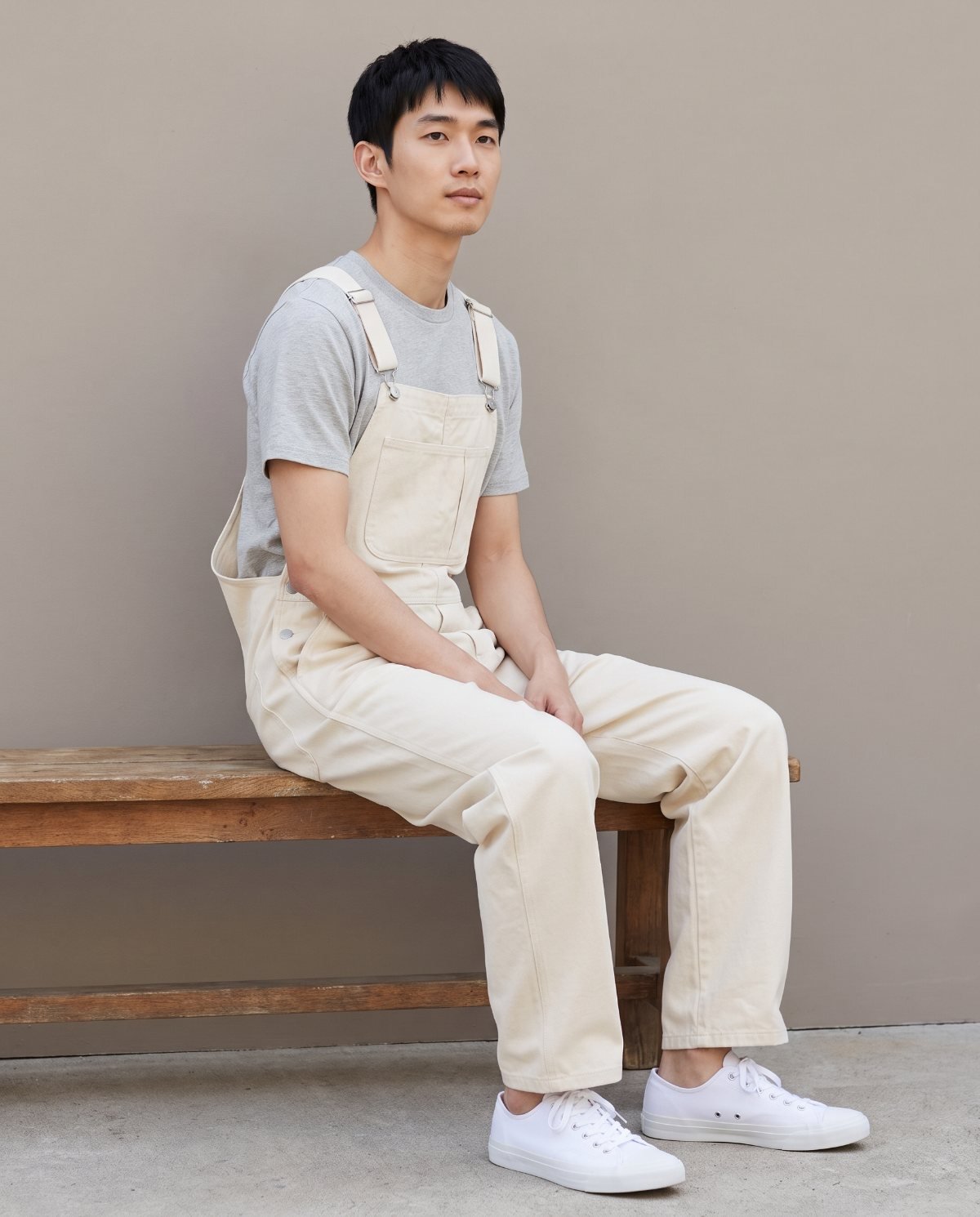 A man seated on a wooden bench in ecru bib overalls and a grey T-shirt, showing how the bib and seat sit when sitting