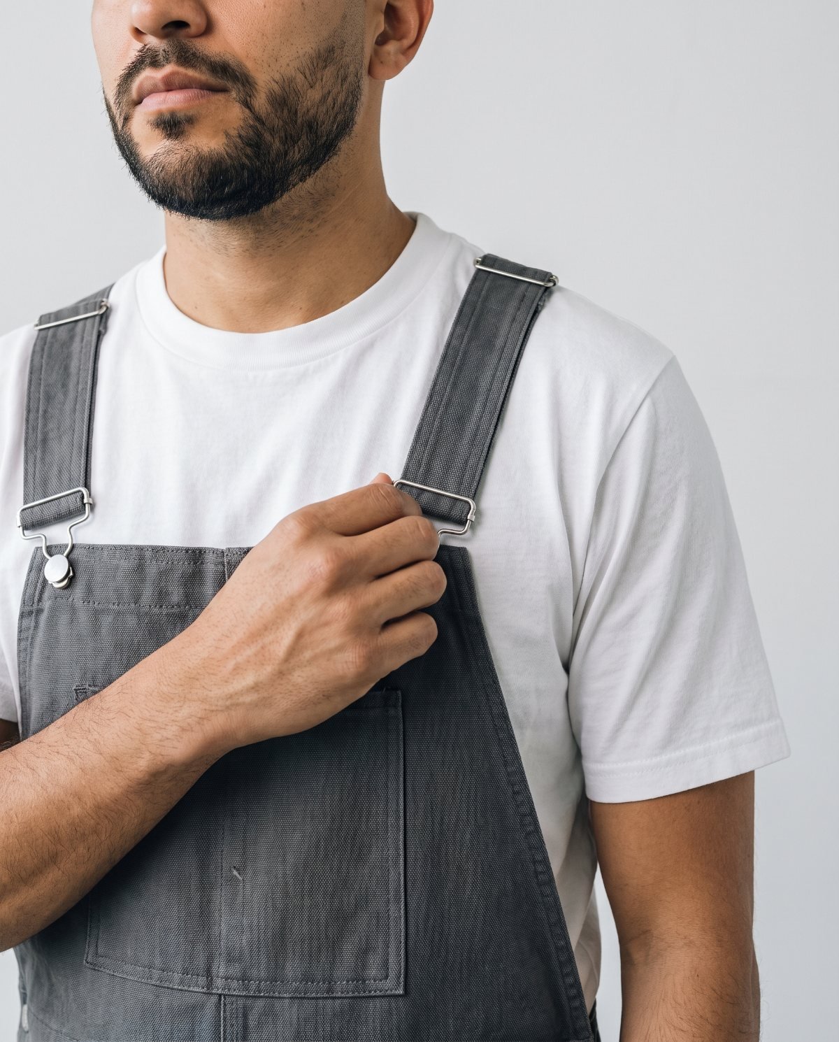 A man adjusting the strap slider of charcoal bib overalls, with the bib sitting at mid-chest over a white T-shirt